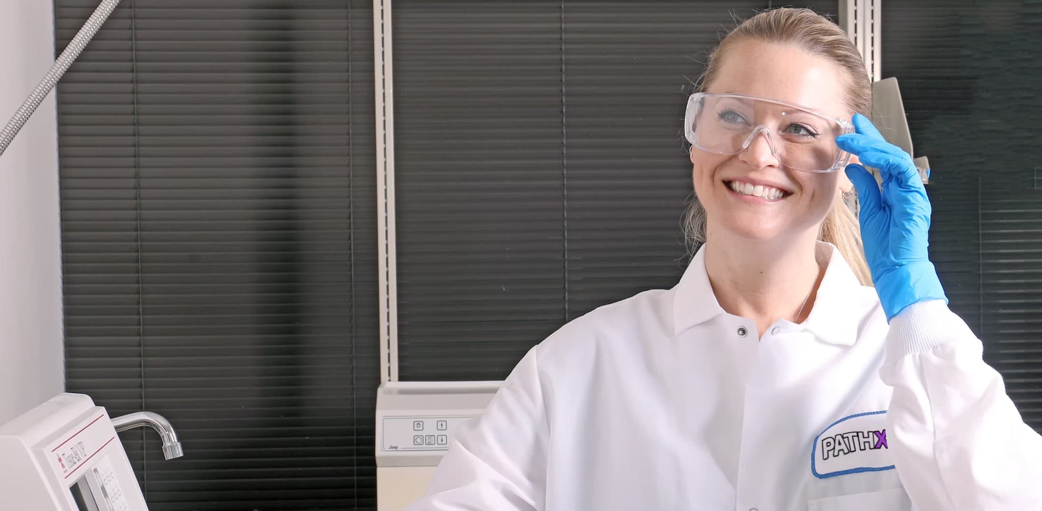 PathXdx laboratory professional in a white coat and safety glasses adjusting protective eyewear beside instrumentation